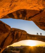 Two mountain bikers ride on an arch in Moab near Lionsback Resort.